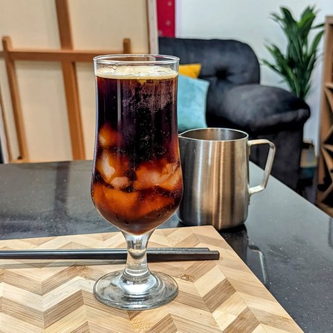 Dark coffee and ice in a tall stemmed glass on a wooden board, with a metal pitcher behind it.