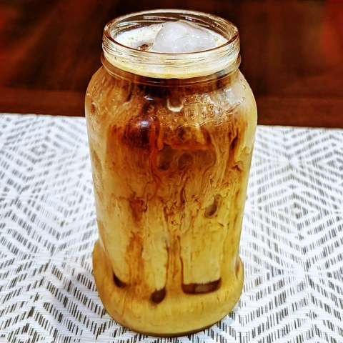 Coffee and milk swirling around ice in a clear jar on a patterned white runner.