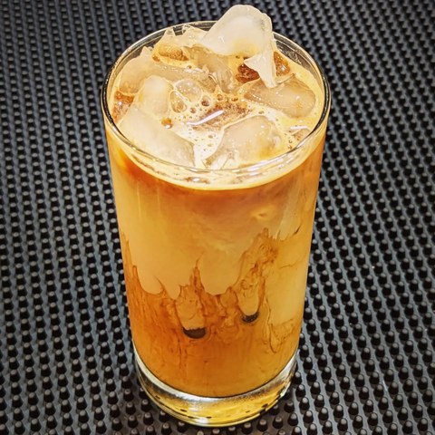Ice and golden swirls of coffee and milk in a tall clear glass on a black bar mat.